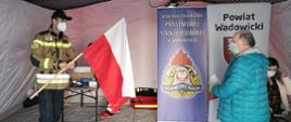 Widok z przodu. W namiocie – mobilnym punkcie szczepień strażak PSP w ubraniu specjalnym wręcza biało-czerwoną flagę pacjentowi, który przystąpił do akcji „Zaszczep się dla Niepodległej”. 
