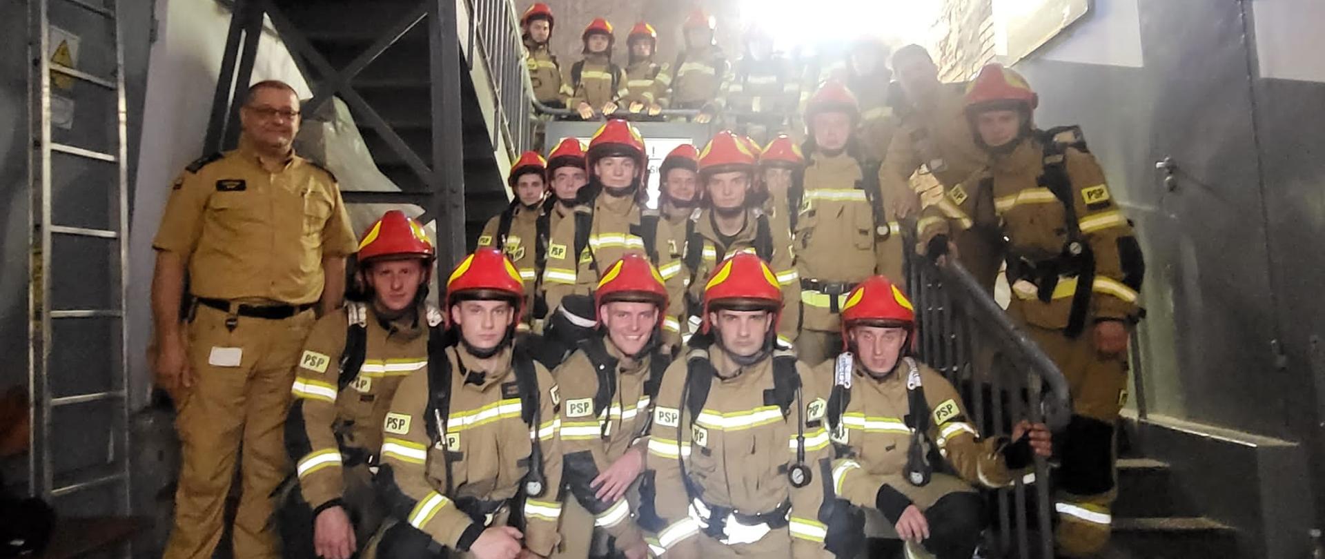 Grupowe zdjęcie kadetów CS PSP w umundurowaniu bojowym z założoną butlą w towarzystwie Zastępcy Komendanta Centralnej Szkoły PSP u podnóża schodów prowadzących na jasnogórską wieżę