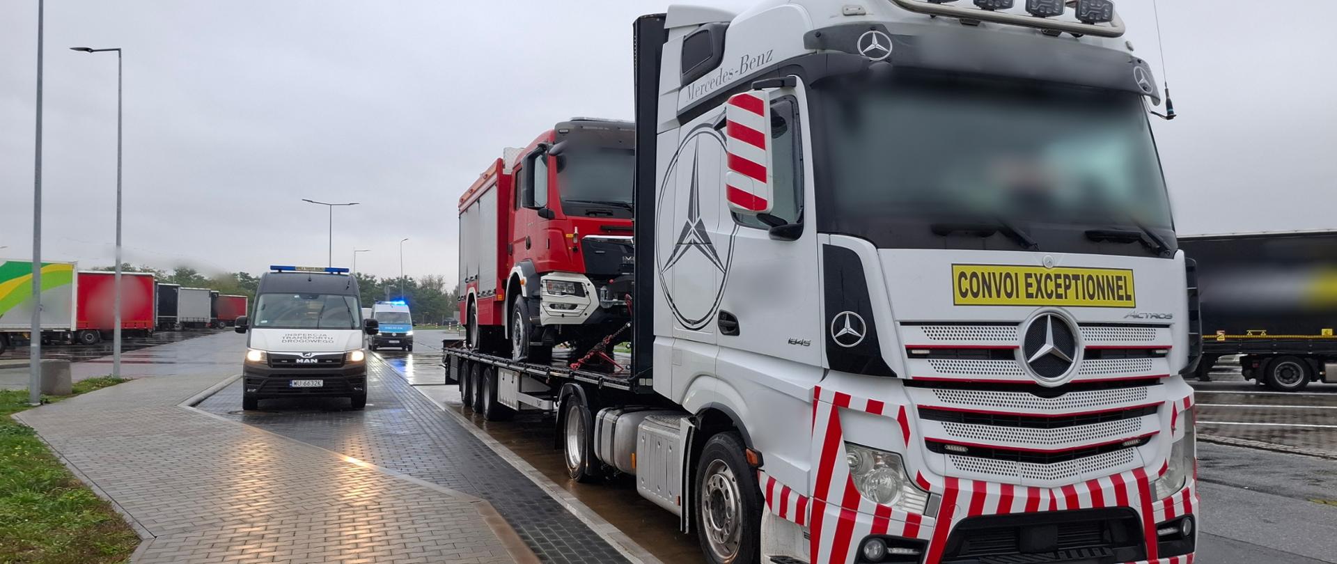 Biały ciągnik siodłowy Mercedes-Benz z oznakowaniem „Convoi Exceptionnel” przewozi na naczepie czerwoną ciężarówkę pożarniczą. Obok stoi oznakowany samochód Inspekcji Transportu Drogowego. Kontrola odbywa się na mokrym parkingu.