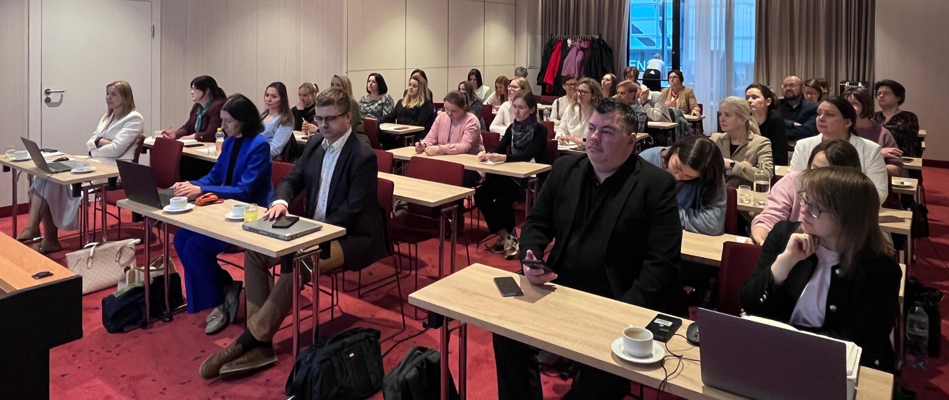 Zdjęcie ukazuje widok na salę konferencyjną wypełnioną uczestnikami. Osoby siedzą przy długich, jasnych stołach ustawionych w rzędach. Większość uczestników to kobiety, niektórzy korzystają z laptopów, robią notatki lub patrzą w stronę przodu sali (niewidocznego na tym ujęciu). Na stołach stoją filiżanki z kawą lub herbatą oraz butelki z wodą. Sala ma nowoczesny wystrój. Pod sufitem zamontowany jest biały projektor multimedialny. W głębi sali, po prawej stronie, widać okno z jasnymi firanami i ciemniejszymi zasłonami.