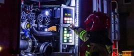 Na zdjęciu widać strażaka operatora-kierowcę znajdującego się przy samochodzie gaśniczym, autopompie. Strażak ubrany w umundurowanie bojowe. Zdjęcie wykonane w nocy. 