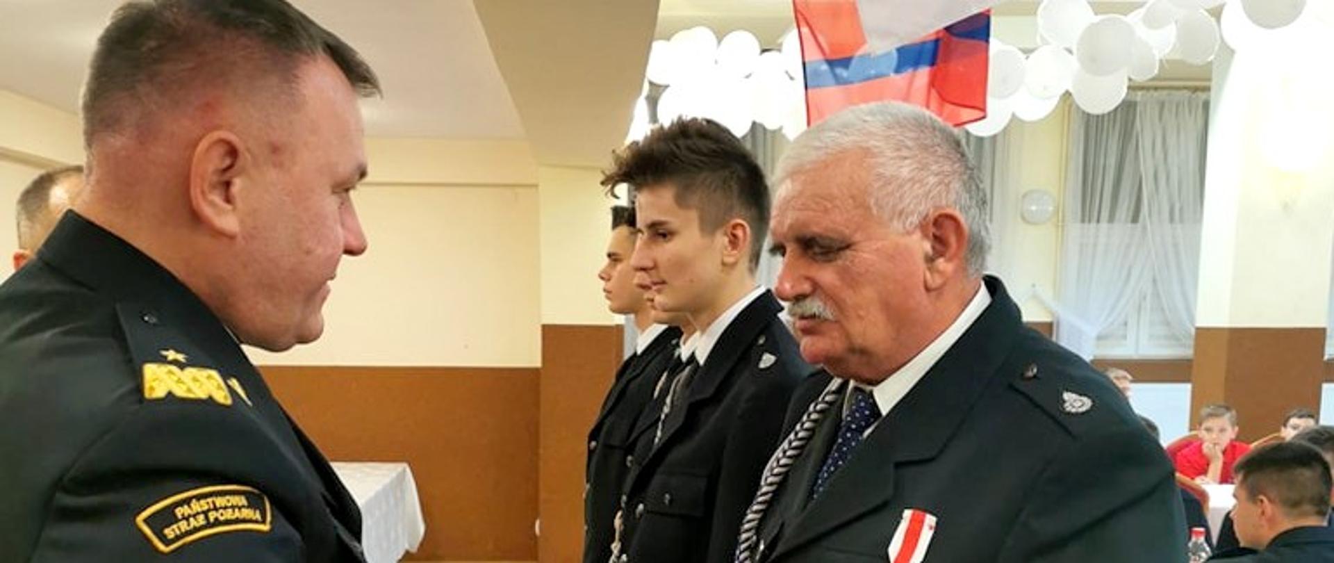 Zdjęcie zrobione w pomieszczeniu, podczas spotkania z druhami ochotnikami. Do zdjęcia pozują ustawieni uczestnicy spotkania, wśród nich strażacy ochotnicy, strażacy Państwowej Straży Pożarnej oraz cywile.