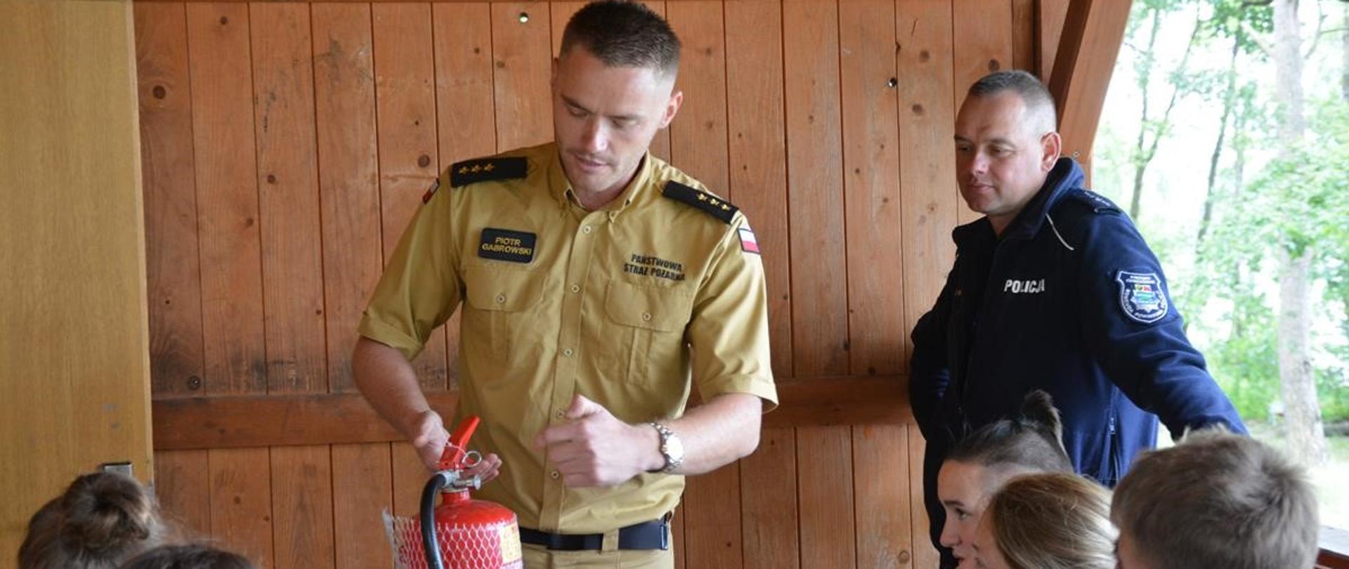 Zdjęcie przedstawia funkcjonariusza Państwowej Straży Pożarnej, który pokazuje uczestnikom wypoczynku zasady obsługi podręcznego sprzętu gaśniczego. 