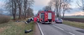 Uszkodzony pojazd w przydrożnym rowie, dookoła porozrzucane elementy pojazdu i bagaże. Na drodze stoi pojazd straży pożarnej i policji.