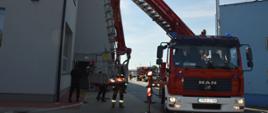 Ćwiczenia taktyczne jednostek ochrony przeciwpożarowej. Teren jednej z firm w Rawiczu. Na pierwszym planie stoi samochód strażacki z podnośnikiem hydraulicznym. W tle inne wozy i strażacy. Z lewej i prawej strony budynki firmy.