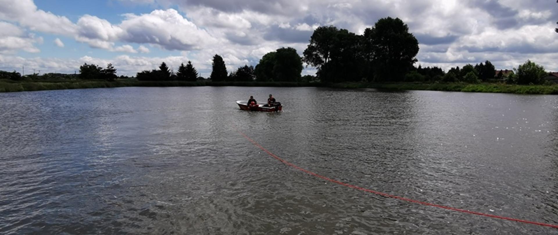 Na łodzi zlokalizowanej na środku stawu znajdują się dwaj strażacy w kamizelkach ratunkowych. Strażacy będą ćwiczyć podejmowanie osób poszkodowanych z wody. W wodzie znajduje się ratownik w ubraniu do pracy w wodzie zabezpieczony linką ratowniczą. Będzie on stymulował osobę poszkodowaną. 