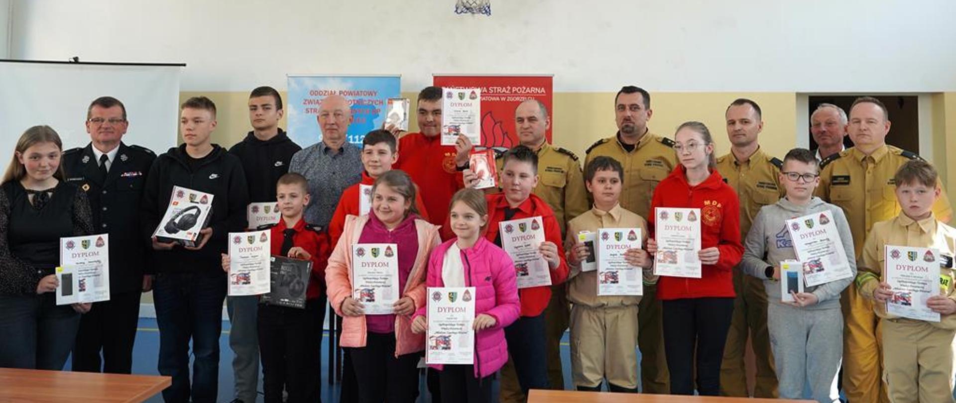 Grupa młodzieży, 4 funkcjonariuszy PSP ubranych w mundury służbowe w kolorze piaskowym, drug OSP w umundurowaniu galowym oraz dwóch mężczyzn w ubraniach cywilnych pozuje do zdjęcia na tle białej ściany. Za pozującymi stoją dwa banery, jedne w kolorze błękitnym, drugi w kolorze czerwonym. Na ścianie wisi tablica do koszykówki.