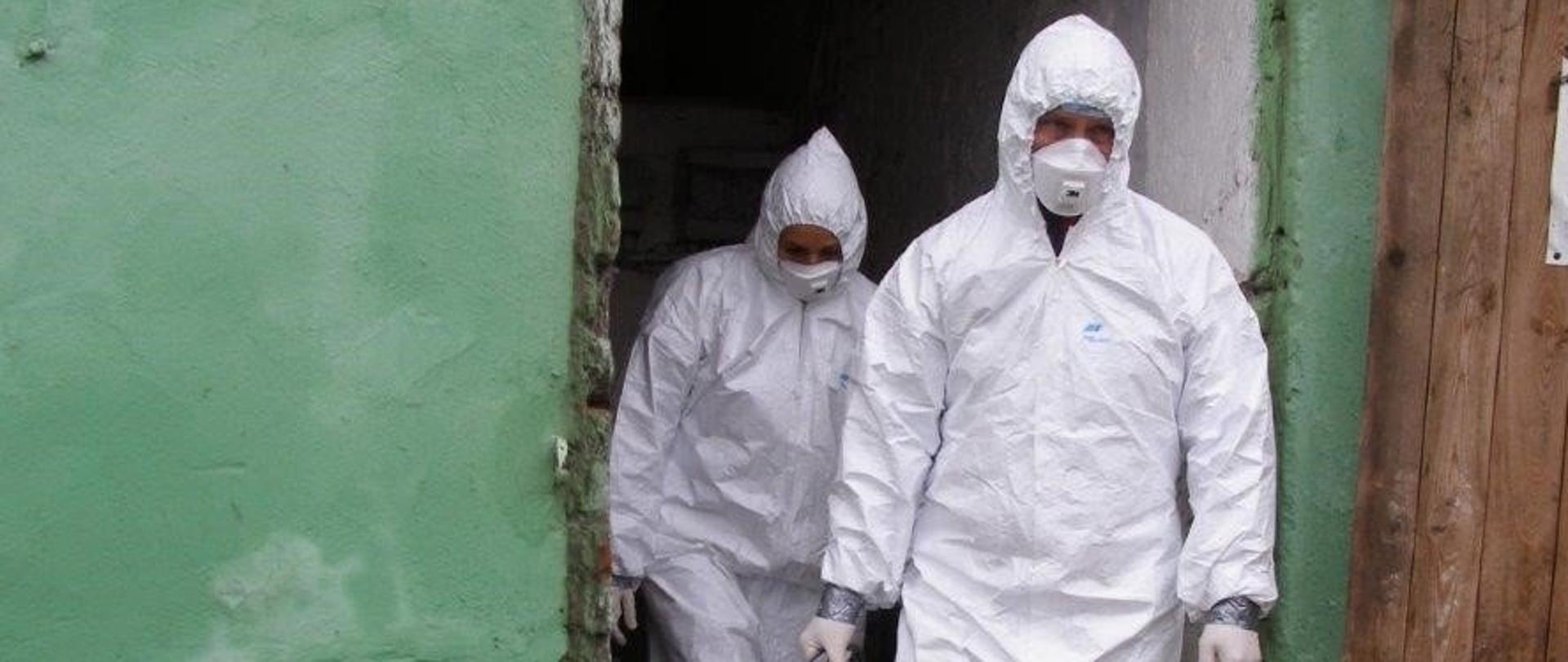 Mężczyźni w strojach zabezpieczających i maseczkach wychodzą z budynku gospodarczego