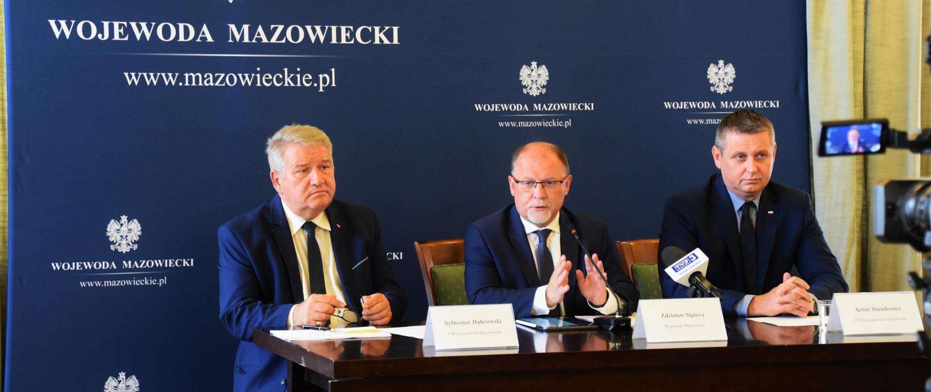 Na pierwszym planie I Wicewojewoda Mazowiecki Sylwester Dąbrowski, Wojewoda Mazowiecki Zdzisław Sipiera i II Wicewojewoda Mazowiecki Artur Standowicz za stołem prezydialnym na tle ścianki wizerunkowej w trakcie konferencji prasowej