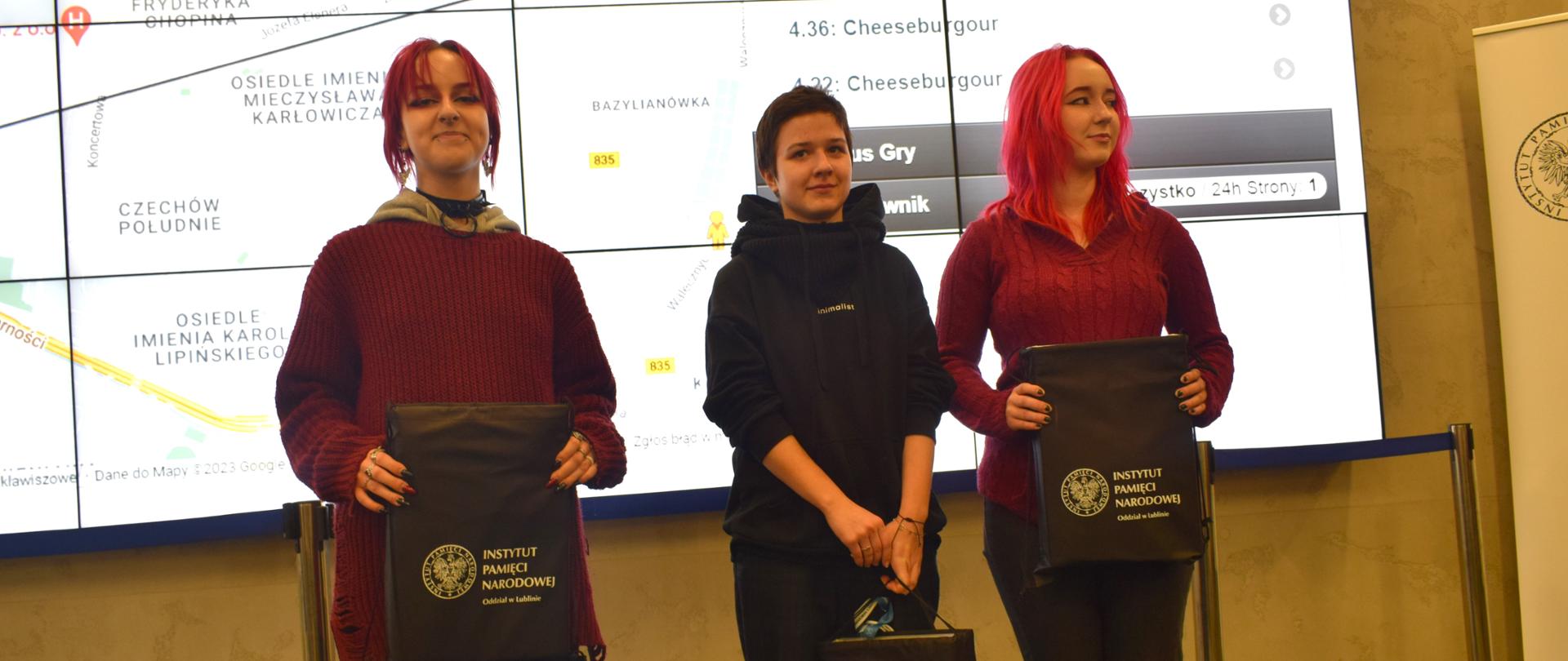 Trzy uczennice w siedzibie IPN w Lublinie stoją z torbami z prezentami z napisem "Instytut Pamięci Narodowej Odział w Lublinie". W tle slajd z fragmentem mapy Google przedstawiająca fragment miasta Lublin i "Widok Gry".
