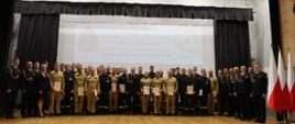 Pamiątkowe zdjęcie absolwentów szkolenia. Na scenie stoją trzymający w rękach teczki z dokumentami uczestnicy wraz z Zastępcą Komendanta Szkoły i pracownikami. 