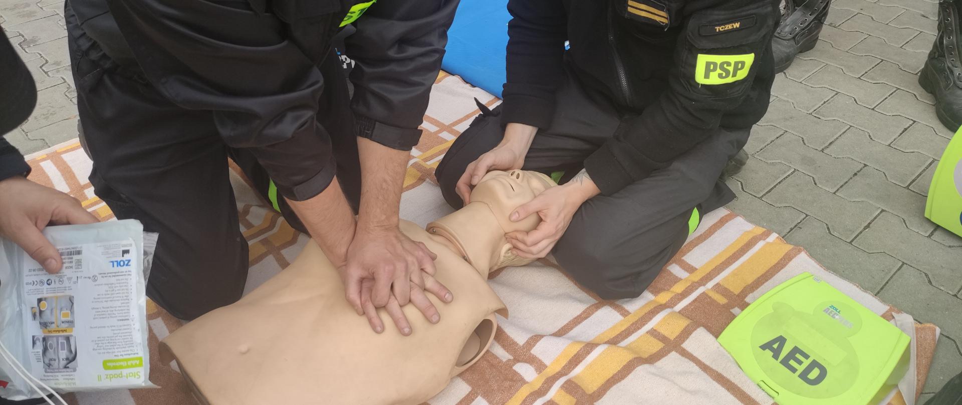 Na kocu leży manekin, którego głowę przytrzymuje strażak PSP. Drugi strażak wykonuje uciski klatki piersiowej. Na zdjęciu również elementy defibrylatora.