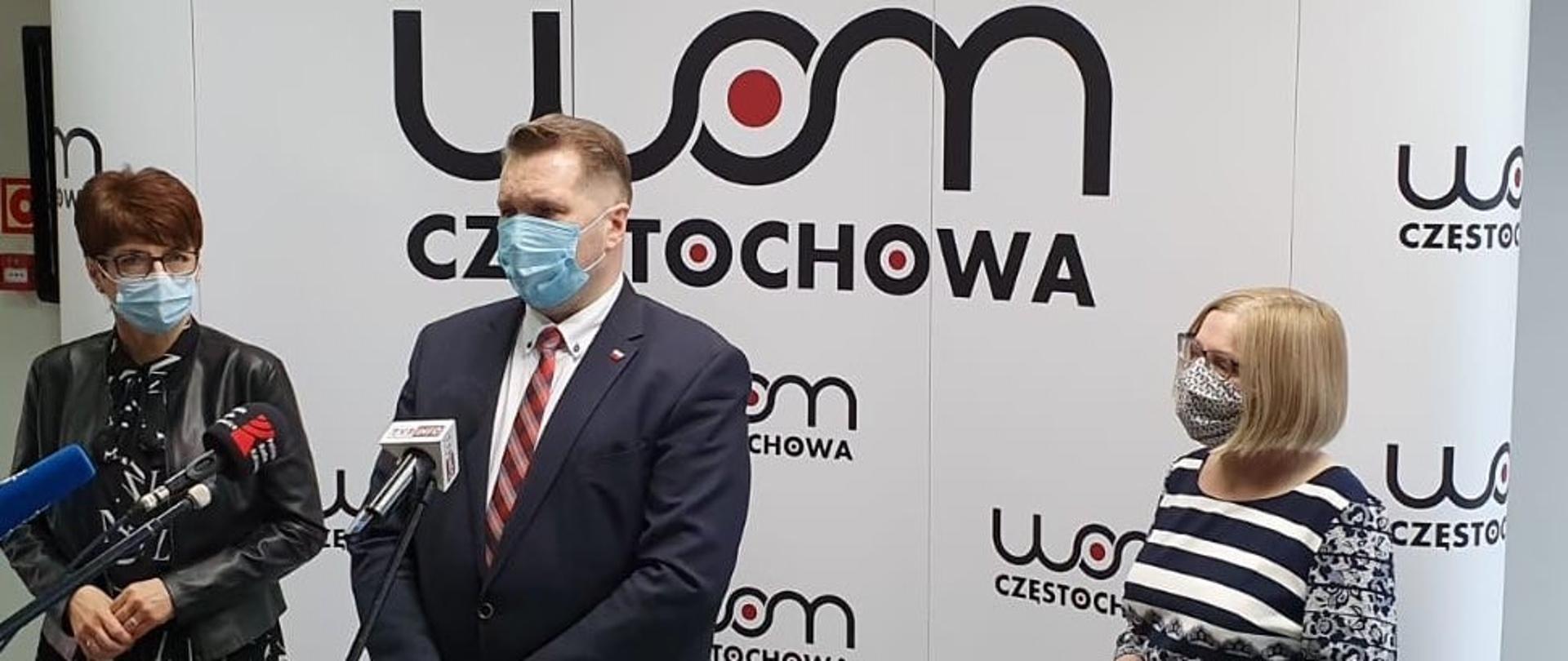 Biała ścianka z logotypem WOM Częstochowa. Na środku zdjęcia minister Przemysław Czarnek stoi przy mikrofonie. Minister ubrany w garnitur ma maseczkę na twarzy. Po lewej stronie ministra Śląski Kurator Oświaty Urszula Bauer. Jest ubrana na ciemno, na twarzy ma okulary i maseczkę. Po prawej stronie kobieta w biało-czarnej sukience. Na twarzy ma okulary i maseczkę. 
