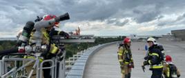 Strażacy stoją na dacu obiektu i przygotowują się do podjęcia osoby poszkodowanej i gaszenia palącej sie aplikacyjnie butli. Do krawędzi dachu przystawiony jest kosz drabiny mechanicznej w którym są strażacy