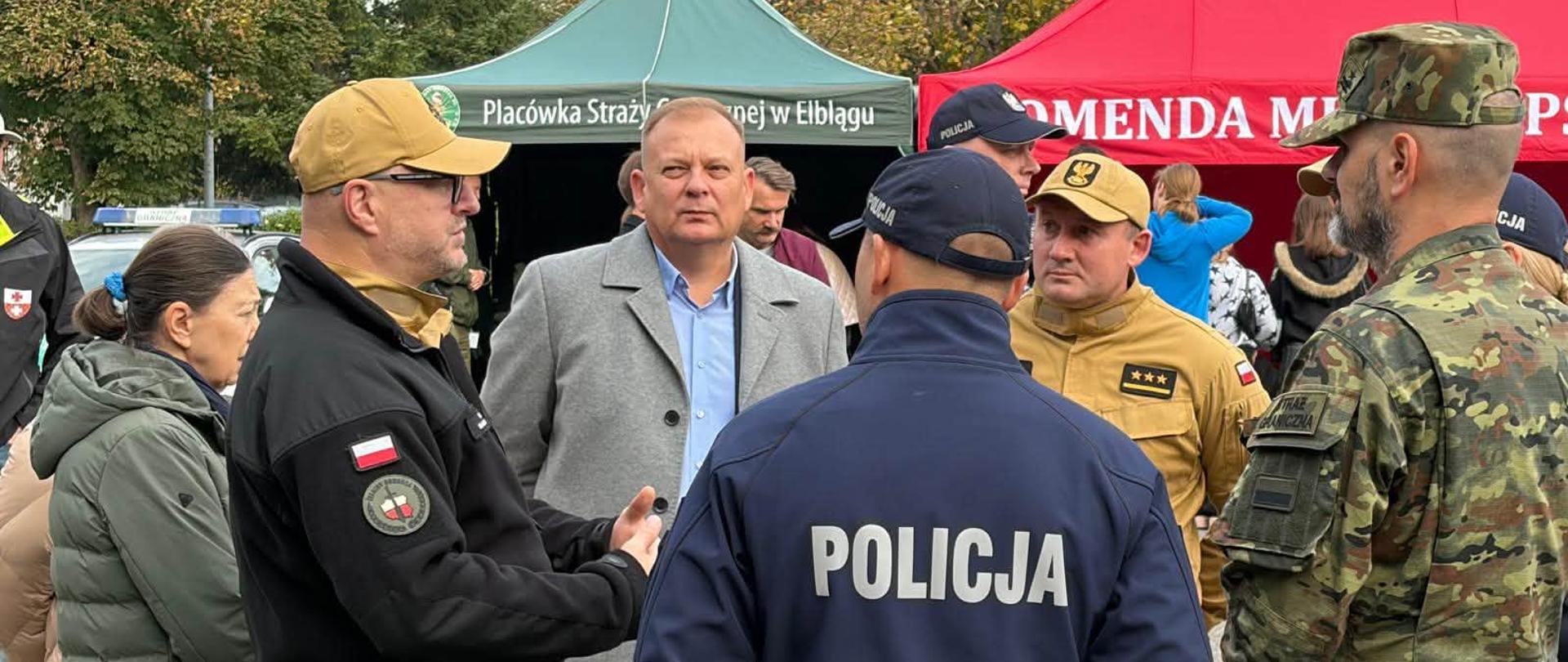 Na pierwszym planie kilka osób prowadzi rozmowę na otwartej przestrzeni podczas wydarzenia plenerowego. Widoczni są przedstawiciele służb mundurowych ubrani w mundury Policji, Państwowej Straży Pożarnej oraz Wojska Polskiego, a także osoby cywilne. W tle znajdują się namioty wystawiennicze, w tym namiot Państwowej Straży Pożarnej w Elblągu oraz namiot Policji. Zdjęcie dokumentuje współpracę i wymianę informacji pomiędzy różnymi służbami odpowiedzialnymi za bezpieczeństwo.