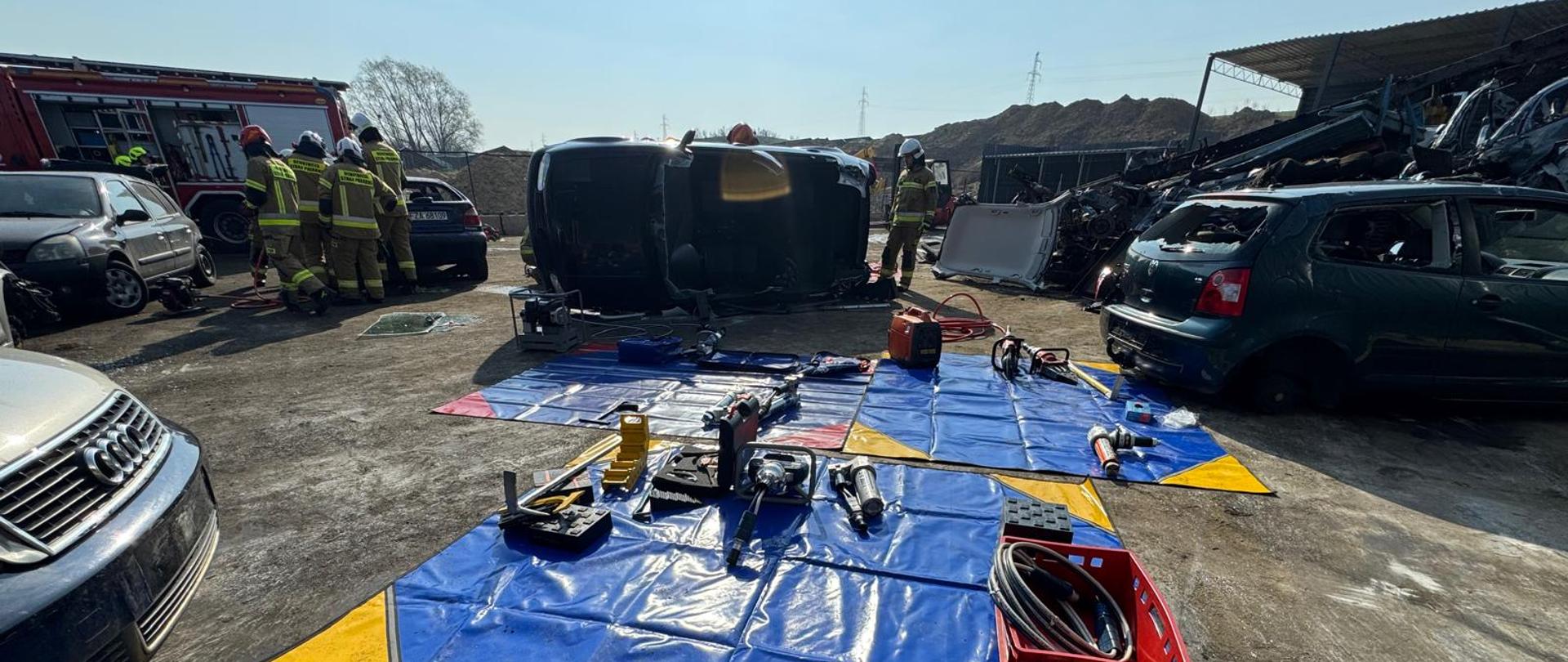 Pole sprzętowe. Sprzęt używany podczas warsztatów znajduje się na plandekach. Za sprzętem znajdują się wraki samochodów oraz przy tych samochodach uczestnicy ćwiczeń.