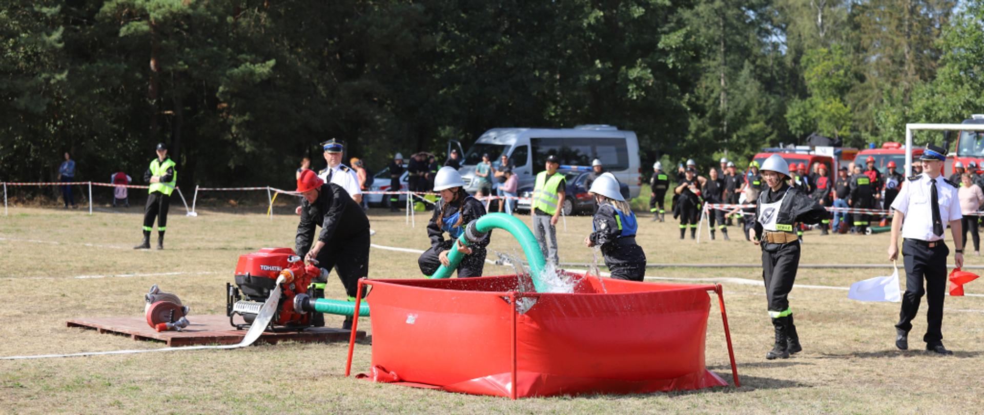 Strażacy podczas wykonywania ćwiczenia bojowego na zawodach sportowo-pożarniczych. Jeden ze strażaków stoi przy pompie i uruchamia ją, drugi strażak wrzuca wąż ssawny do zbiornika z wodą, trzeci strażak biegnie w stronę podestu, czwarty strażak stoi z tyłu i obserwuje. Strażacy ubrani w hełmy i ubrania koszarowe. Całość ćwiczenia oceniają stojący w pobliżu funkcjonariusze – sędziowie. Sędziowie ubrani w koszule krótkie z pagonami, krawatem oraz rogatywki. W tle widać samochody pożarnicze, strażaków w ubraniach koszarowych oraz las. Zdjęcie wykonano w słoneczny dzień na boisku w Porytem-Jabłoni.