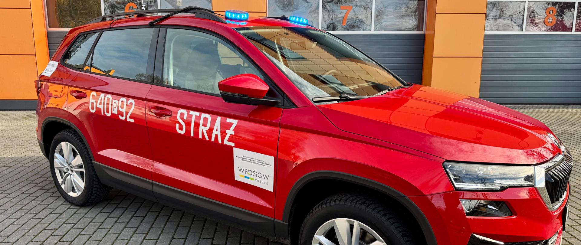 Na zdjęciu widać czerwony samochód typu SUV oznaczony jako pojazd strażacki. Na bokach auta białymi literami widnieje napis „STRAŻ” oraz numer operacyjny 640[P]92. Samochód ma zamontowane niebieskie światła sygnalizacyjne na dachu. Na przednich drzwiach znajduje się również naklejka z logo instytucji wspierającej zakup pojazdu WFOŚiGW. Auto stoi na brukowanej powierzchni, w tle widać ogrodzenie boiska sportowego oraz budynki. 