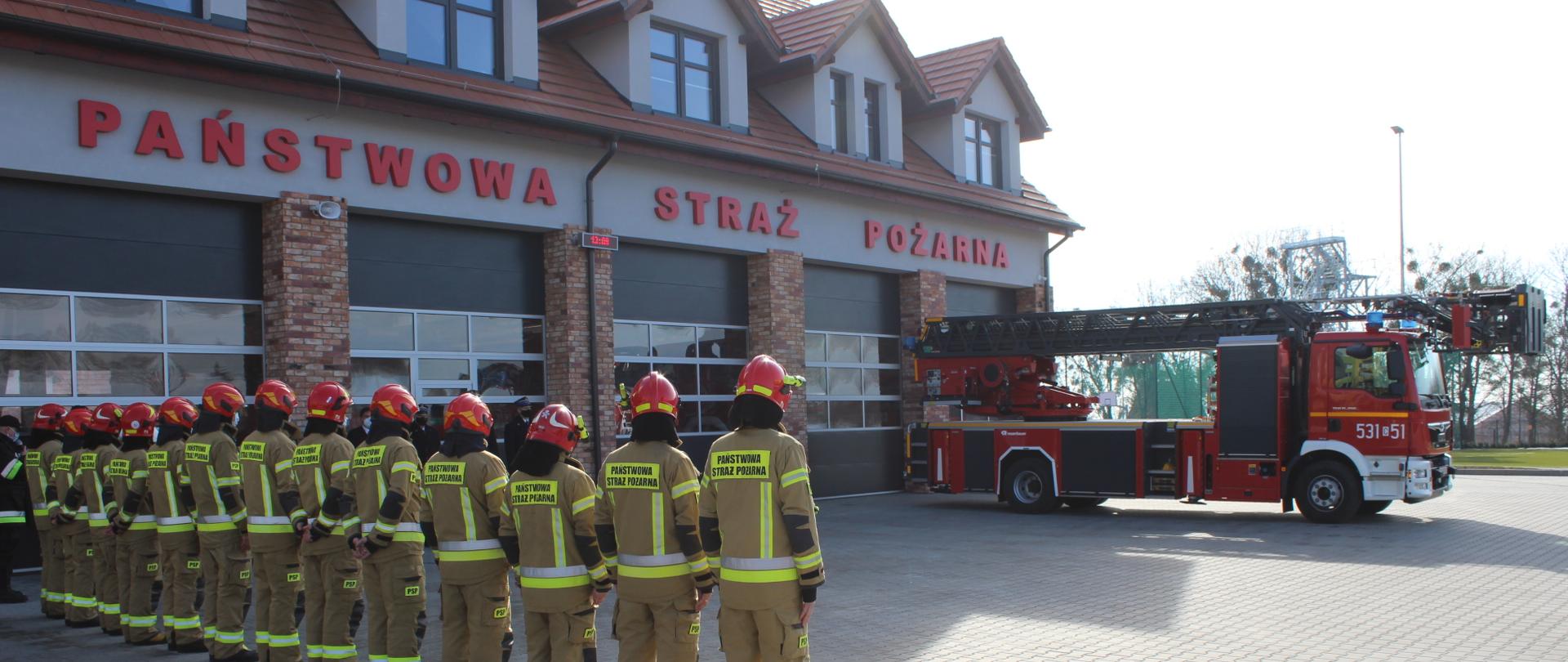 Na środku zdjęcia stoi Dyrektor Generalny Rosenbauer Karlsruhe Pana Michael Kristeller z założoną maseczką, przemawia do mikrofonu, na tle nowego samochodu pożarniczego z drabiną mechaniczną.