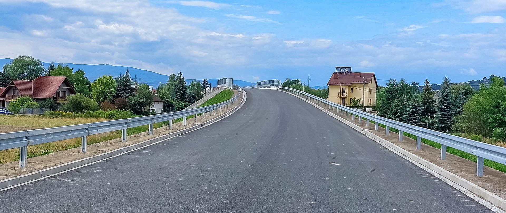 Fragment obwodnicy Chełmca. Wznosząca się jezdnia z nowym asfaltem. Po lewej i prawej stronie barierki. W oddali widoczne zabudowania, góry i drzewa.