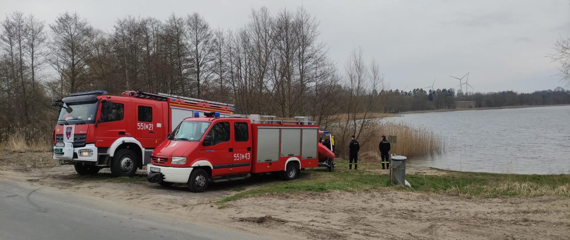 Zdjęcie przedstawia jezioro oraz brzeg na którym znajduja sie dwa wozy strażackie.