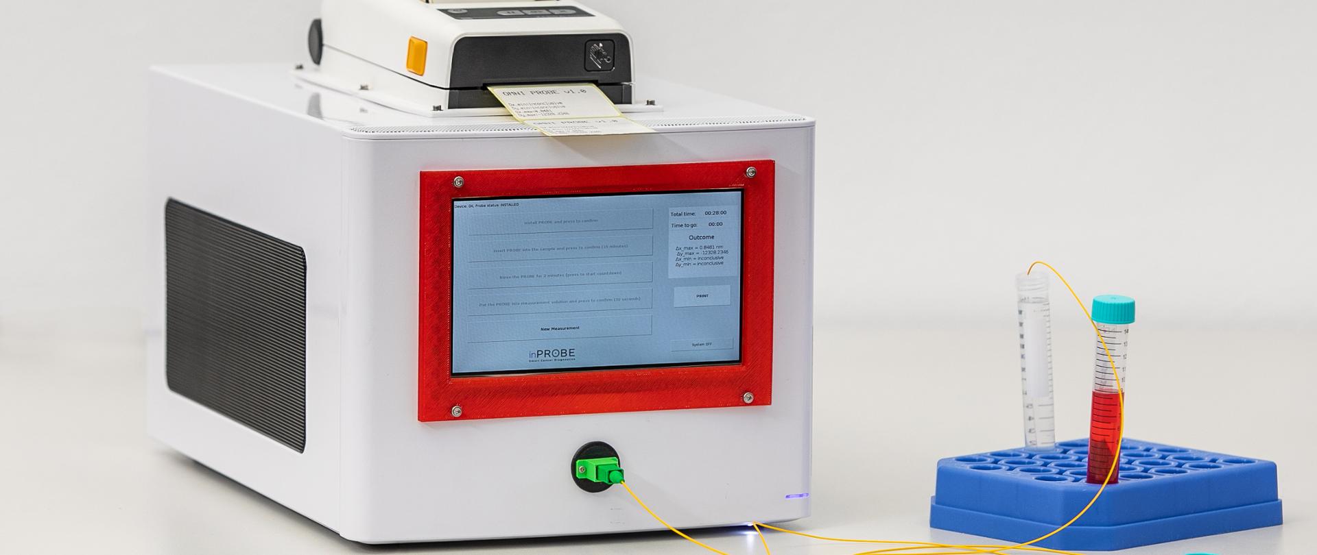 Urządzenie diagnostyczne w kształcie białej skrzynki w metalowej obudowie, z ekranem i możliwością wydruku wyników. Urządzenie jest połączone z probówką za pomocą cienkich przewodów.