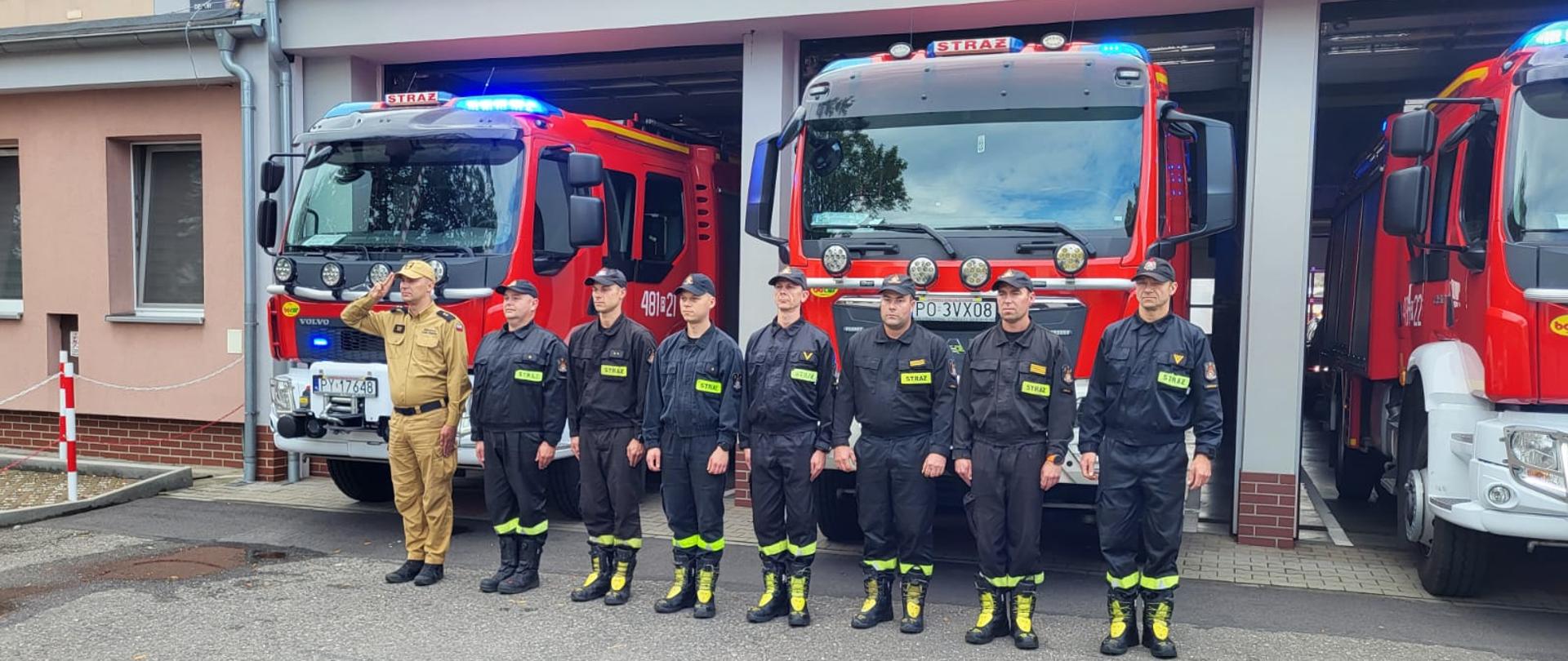 Strażacy oddają hołd Powstańcom Warszawy. Na zdjęciu widać strażaków przed budynkiem Komendy, samochody strażackie wyjechały z garażu, mają włączone sygnały świetlne i dźwiękowe, widać maszt z flagą Polski i budynek Komendy Powiatowej PSP w Kościanie.