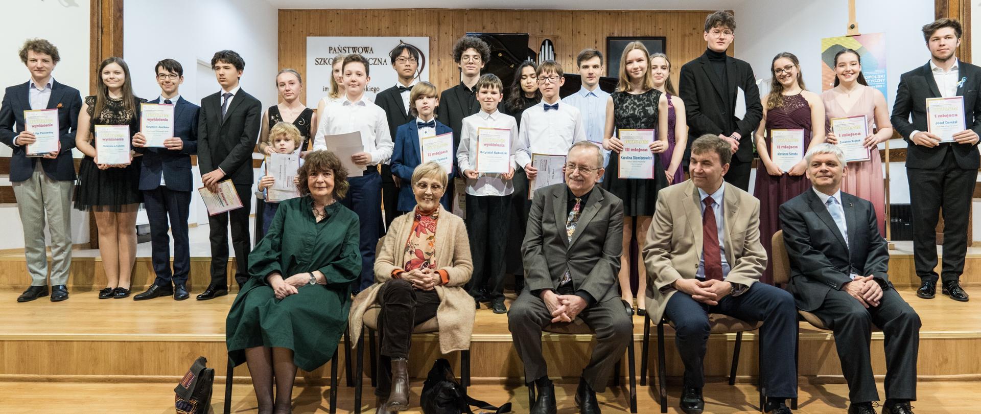 Sala koncertowa PSM w Koninie, jury i dyrektor szkoły siedzą na krzesłach (5 osób), laureaci stoją w dwóch rzędach 21 osób ubranych na galowo.