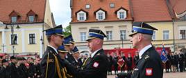 Na pierwszym planie oficer w ciemnym mundurze galowym przypina medal do piersi strażaka stojącego przed nim na baczność. Obok nich stoi trzeci funkcjonariusz, trzymający etui z kolejnymi odznaczeniami. W tle widoczny jest szereg młodych strażaków w czarnych mundurach koszarowych, poczty sztandarowe oraz zabytkowe kamienice o jasnych elewacjach.