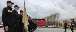 Na zdjęciu widzimy strażaków ustawionych w szyku w czarnych czapkach z daszkiem, czarnych mundurach, pasach głównych z toporkami oraz w czarnych butach typu bukaty podkute. Pododdział ćwiczy elementy uroczystości pożarniczych. Na zdjęciu widzimy również przed pododdziałem osoby szkolące również w mundurach strażackich.
