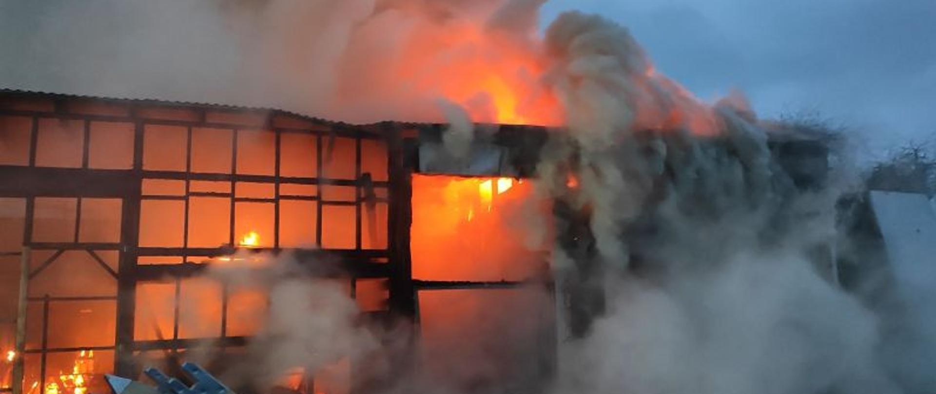 Jezdnia brukowa, cztery duże samochody pożarnicze z pootwieranymi skrytkami i włączonymi światłami alarmowymi. Przy jednym stoi strażak. W oddali jeszcze dwóch strażaków.