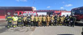 Zdjęcie przedstawia dużą grupę strażaków ustawioną w szeregu przed budynkiem Jednostki Ratowniczo-Gaśniczej. W tle widoczne są trzy wozy strażackie oraz czerwona brama garażowa. Strażacy ubrani są w różne rodzaje ubrań ochronnych – zarówno PSP, jak i OSP. Na niebie widoczne jest słońce i kilka chmur, co świadczy o dobrej pogodzie. Zdjęcie zostało wykonane prawdopodobnie po zakończeniu ćwiczeń lub odprawy.