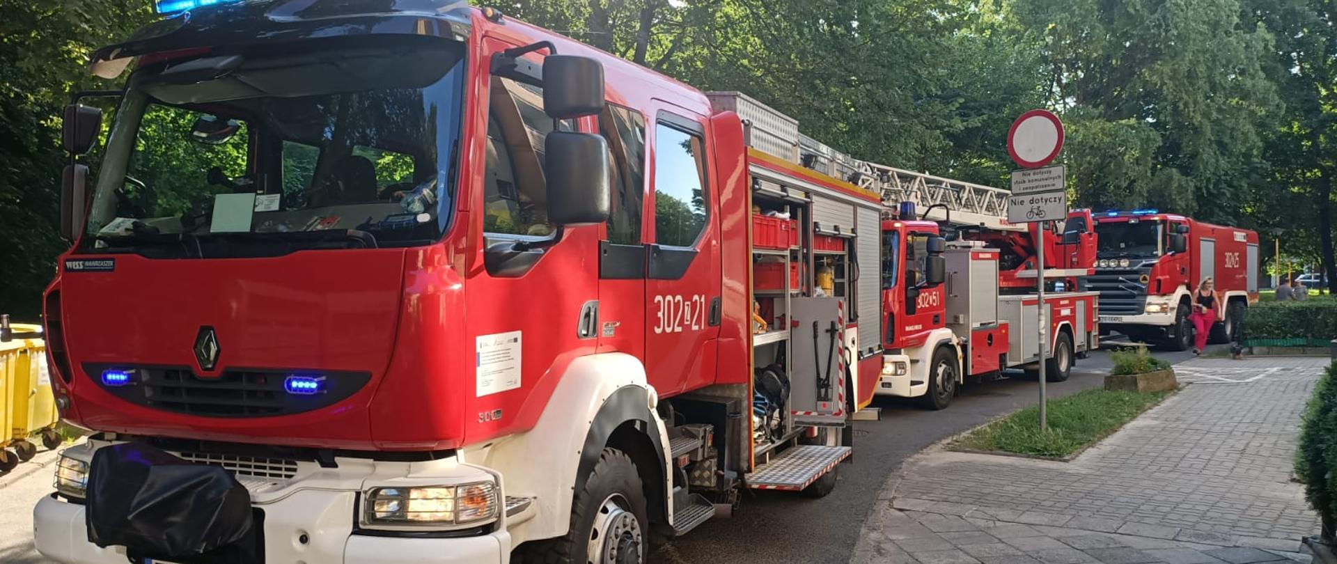 Na zdjęciu widzimy trzy wozy bojowe uczestniczące w zdarzeniu stojące na drodze pod blokiem. Zdjęcie zrobione od przodu pojazdów stojących na sygnałach świetnych.