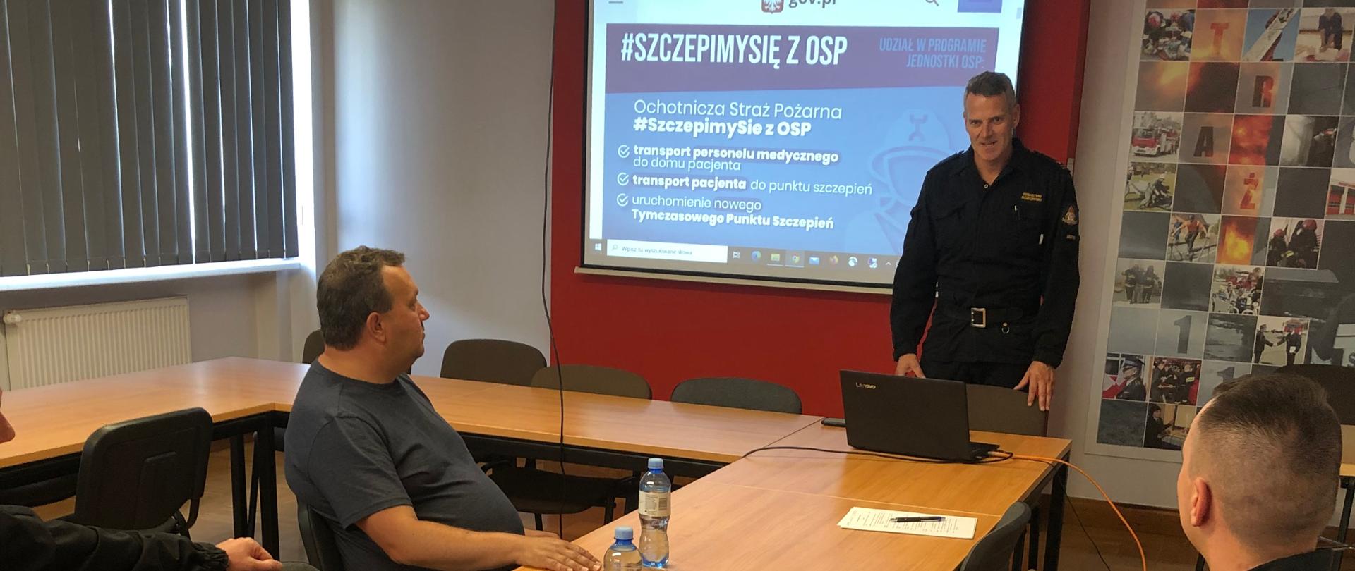 Strażak stoi przemawiając do siedzących strażaków i osób cywilnych. W tle ekran z wyświetloną prezentacją.