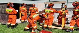 Zdjęcie przedstawia grupę strażaków biorących udział w ćwiczeniach, są oni ubrani w specjalne pomarańczowe ubrania wraz z kaskiem. Na pierwszym planie widzimy strażaków przygotowujących podporę do montażu w celu