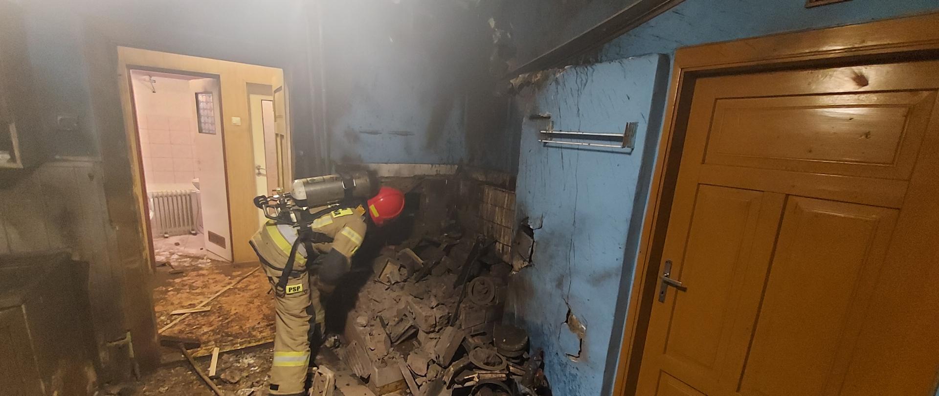 Zdjęcie przedstawia strażaka w żółtym ubraniu sprawdzającego gruzy po wybuchu kuchni kaflowej