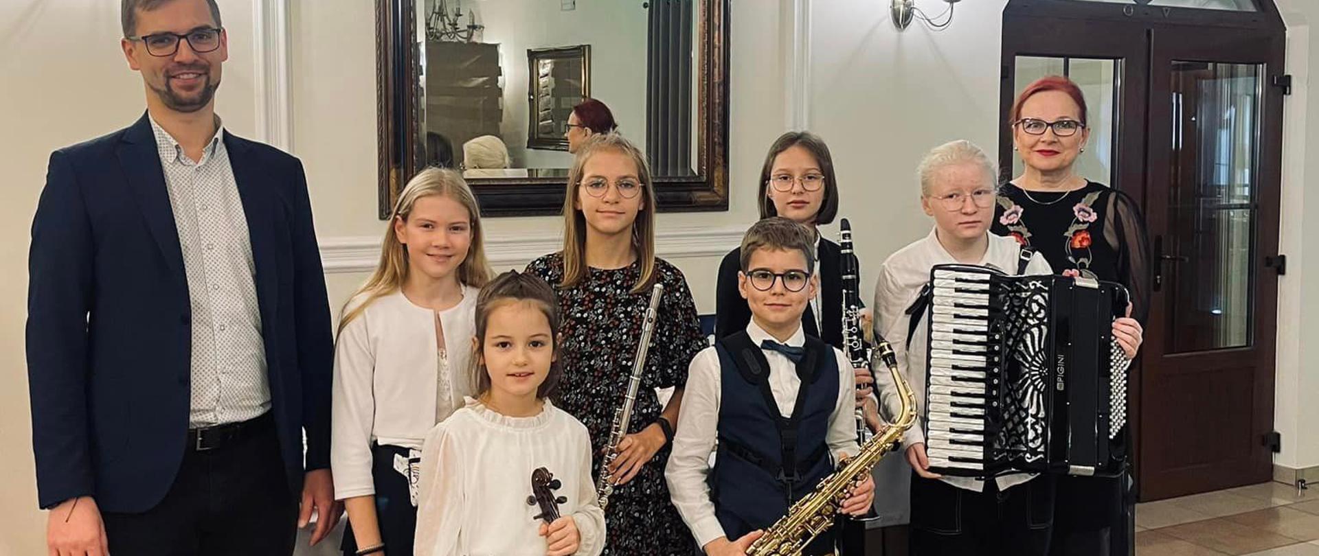 Grupa uczniów wraz z nauczycielami w hallu budynku, trzymają saksofon, skrzypce, flet, akordeon i klarnet