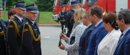 Na zdjęciu widzimy funkcjonariuszy, którzy wręczają zaproszonym gościom okolicznościowe statuetki. Strażacy ubrani są w mundury galowe i czarne rękawiczki. Osoby odbierające statuetki ubrani są w garnitury mężczyźni) kobiety ubrane w płaszcze.
