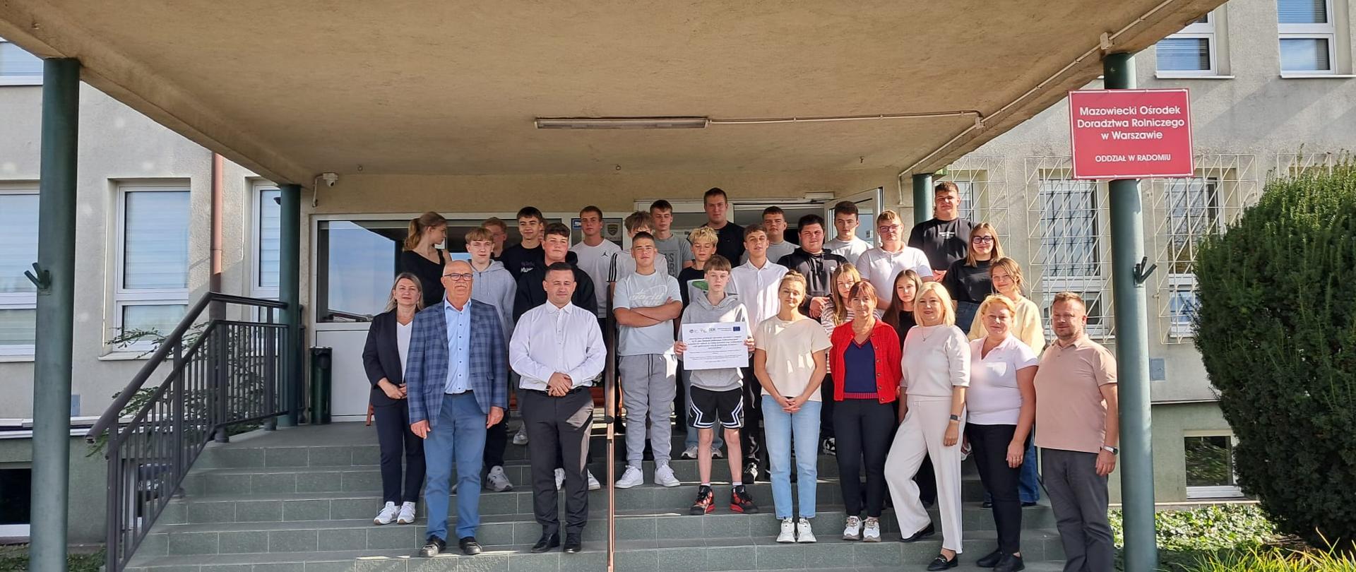 Kilkudziesięcioosobowa grupa uczniów i dorosłych stoi na schodach przed wejściem do budynku z napisem Mazowiecki Ośrodek Doradztwa Rolniczego w Warszawie Oddział w Radomiu. Uczeń w pierwszym rzędzie trzyma tablicę informacyjną.