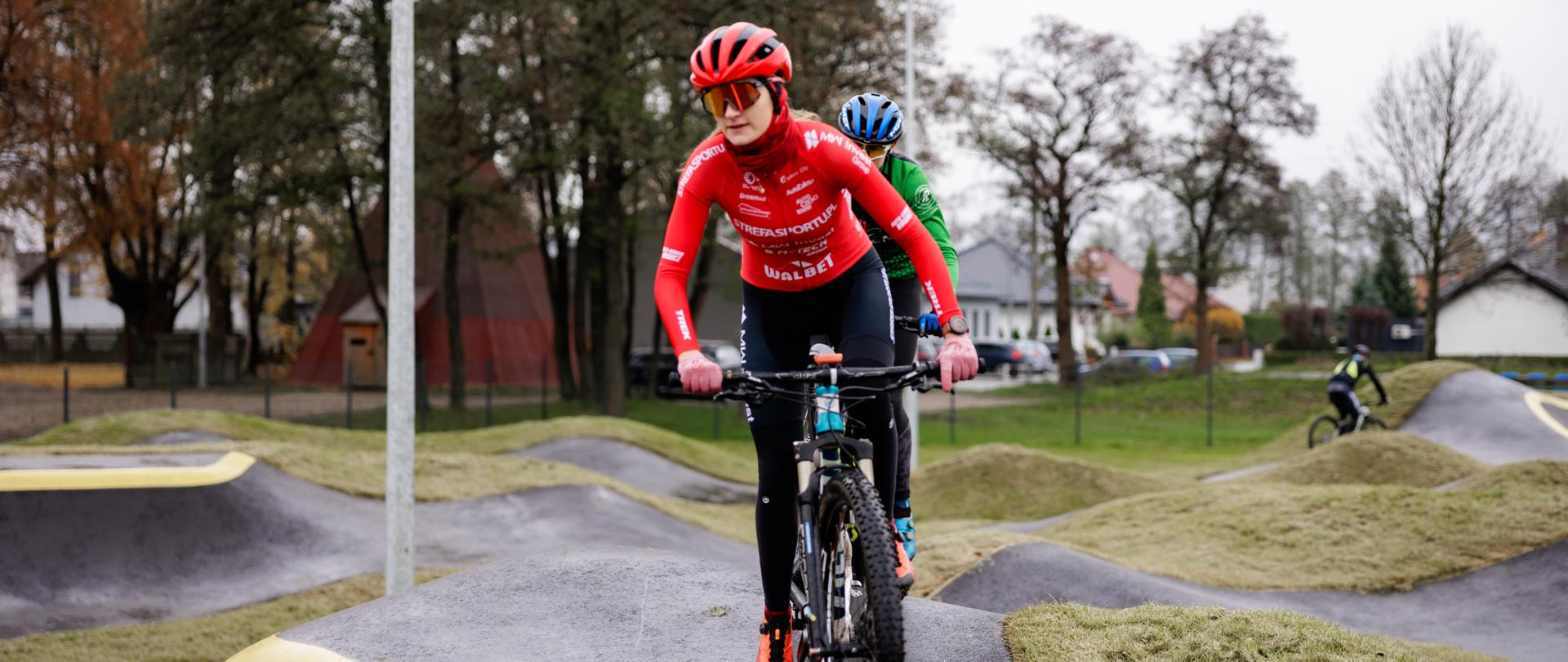 Zdjęcie przedstawiające dwójkę ludzi jeżdżących na rowerach po pumptracku. Z tyłu widać trzecią osobę. W tle drzewa i zabudowania.