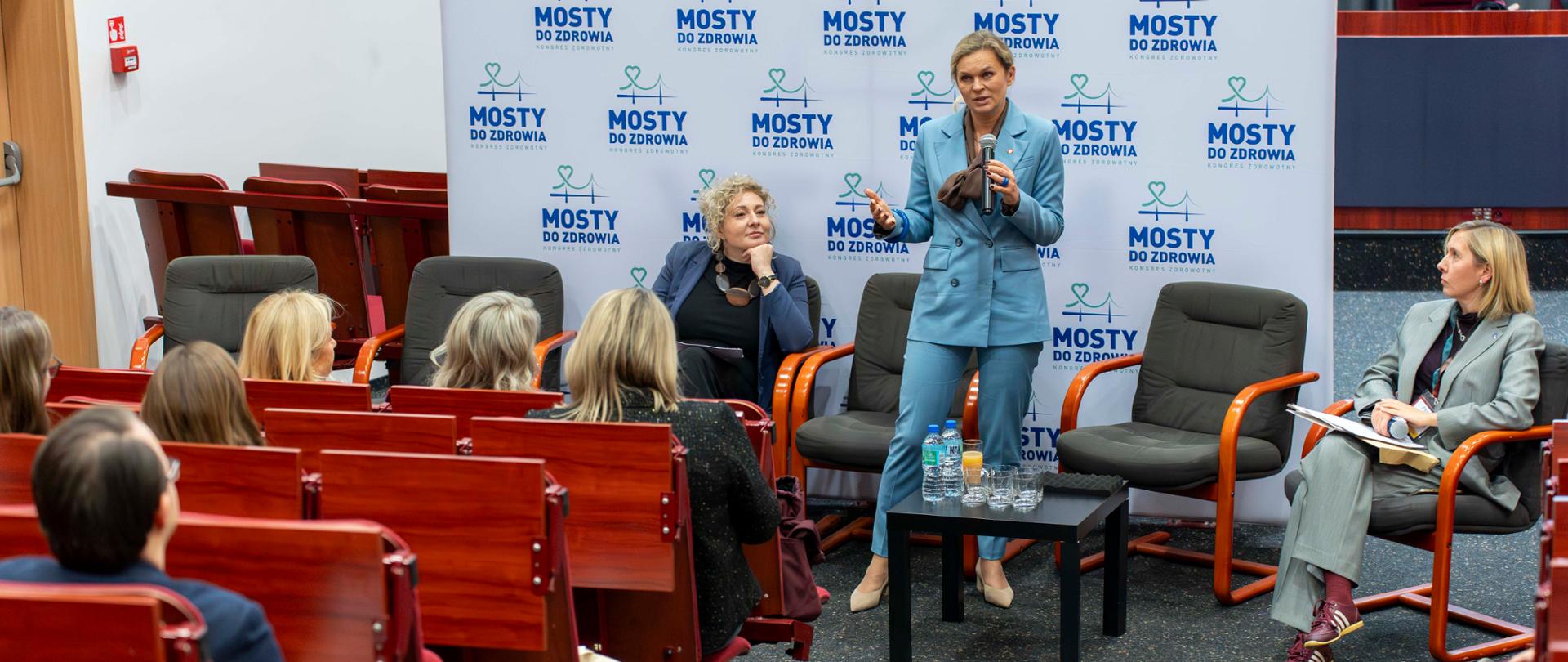 Szerokie ujęcie sali konferencyjnej. Na scenie stoi Barbara Nowacka, minister edukacji, w jasnoniebieskim garniturze, przemawiając do mikrofonu. Obok niej na krzesłach siedzą dwie inne panelistki. Na pierwszym planie widoczni są uczestnicy konferencji siedzący w drewnianych rzędach krzeseł, słuchający prelegentki. W tle ściana z logo konferencji "MOSTY DO ZDROWIA".