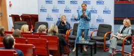 Szerokie ujęcie sali konferencyjnej. Na scenie stoi Barbara Nowacka, minister edukacji, w jasnoniebieskim garniturze, przemawiając do mikrofonu. Obok niej na krzesłach siedzą dwie inne panelistki. Na pierwszym planie widoczni są uczestnicy konferencji siedzący w drewnianych rzędach krzeseł, słuchający prelegentki. W tle ściana z logo konferencji "MOSTY DO ZDROWIA".