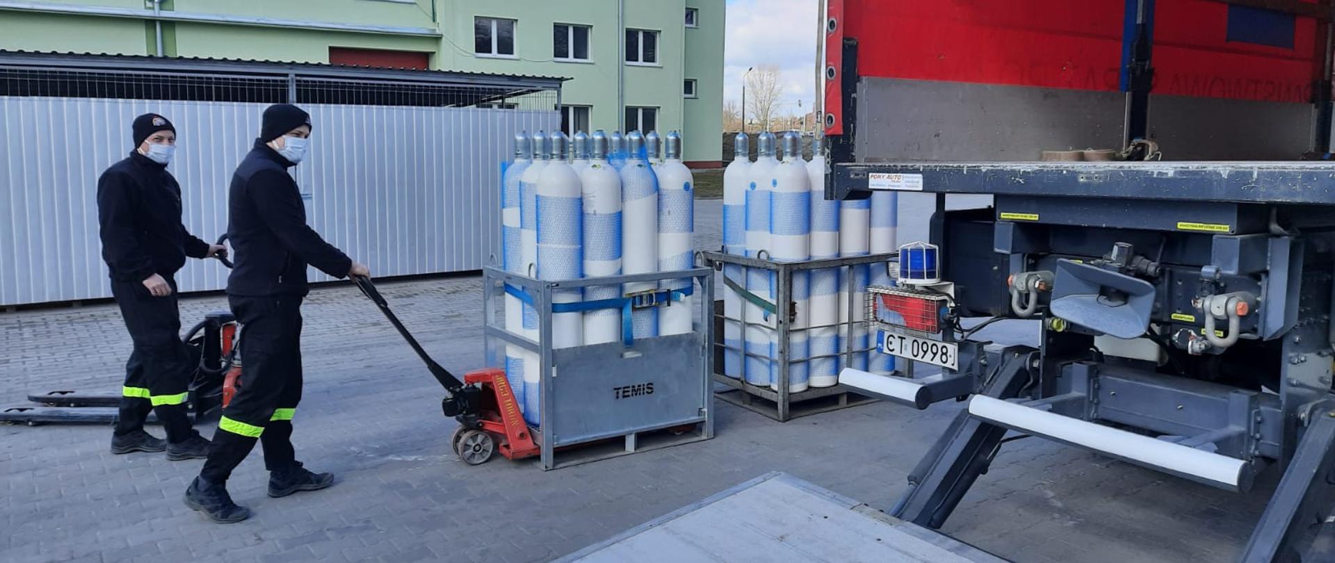Strażacy podczas przygotowywania transportu butli z tlenem do Szpitala SP ZOZ Łasin. Butle z tlenem zapakowane w kosze transportowe są przewożone przy pomocy wózków paletowych i umieszczane w samochodzie kwatermistrzowskim widocznym z prawej strony. W tle widać wiaty banku tlenu oraz budynek Jednostki Ratowniczo - Gaśniczej nr 3 w Toruniu.