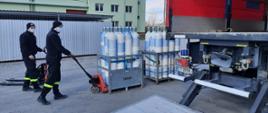 Strażacy podczas przygotowywania transportu butli z tlenem do Szpitala SP ZOZ Łasin. Butle z tlenem zapakowane w kosze transportowe są przewożone przy pomocy wózków paletowych i umieszczane w samochodzie kwatermistrzowskim widocznym z prawej strony. W tle widać wiaty banku tlenu oraz budynek Jednostki Ratowniczo - Gaśniczej nr 3 w Toruniu.