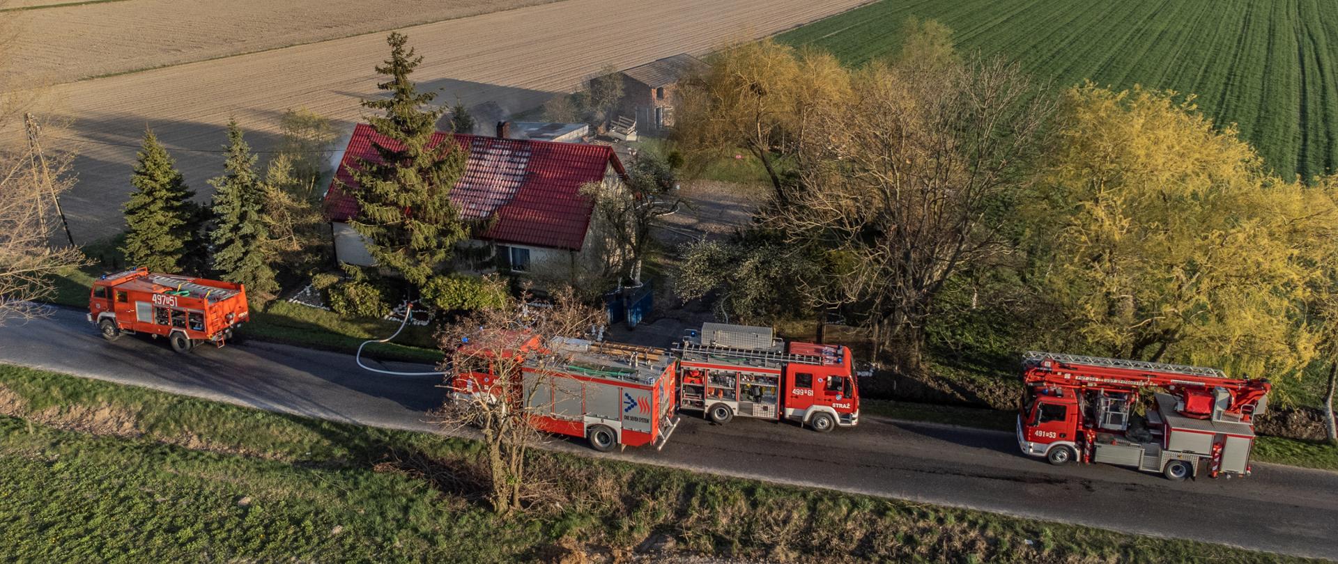 Na drodze przebiegającej w pobliżu budynku mieszkalnego stoją pojazdy pożarnicze. Nad dachem unosi się dym. W oddali widać strażaków prowadzących działania na terenie posesji. Pokrycie dachowe w miejscu pożaru zmieniło barwę. Zdjęcie wykonane z drona,