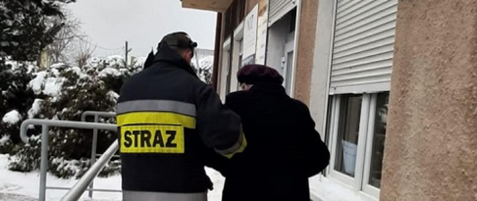 Na zdjęciu widać strażaka, który pomaga wsiąść do samochodu służbowego starszej kobiecie. Zaśnieżony chodnik. W oddali widać kawałek płotu, drzewo iglaste i blok mieszkalny.