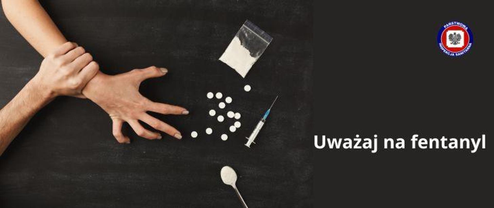 Na ciemnym ręka sięgająca po substancje psychoaktywne białe tabletki, strzykawkę i biały proszek w foliowej torebce. Druga ręka zaciśnięta na pierwszej w celu powstrzymania przed użycia narkotyków. Po prawej stronie biały napis uważaj na fentanyl. W prawy górnym rogu logo Państwowej Inspekcji Sanitarnej.