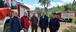 Zdjęcie wykonano w ciągu dnia podczas manewrów Krajeńskie Niwy 2022. Komendant Powiatowy ubrany w mundur służbowy oraz nadleśniczy pozuje do zdjęcia z gośćmi na tle samochodów ratowniczo-gaśniczych.