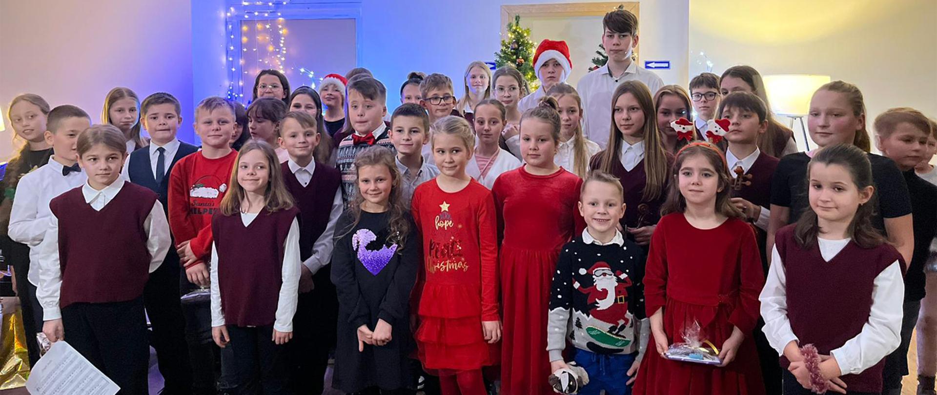 Duża grupa uczniów stoi w auli szkoły, za nimi widać udekorowaną choinkę, dzieci ubrani są w bożonarodzeniowe stroje.