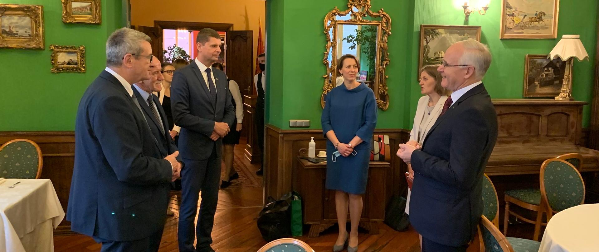 Minister i członkowie delegacji stoją w pokoju, dookoła zabytkowe meble.